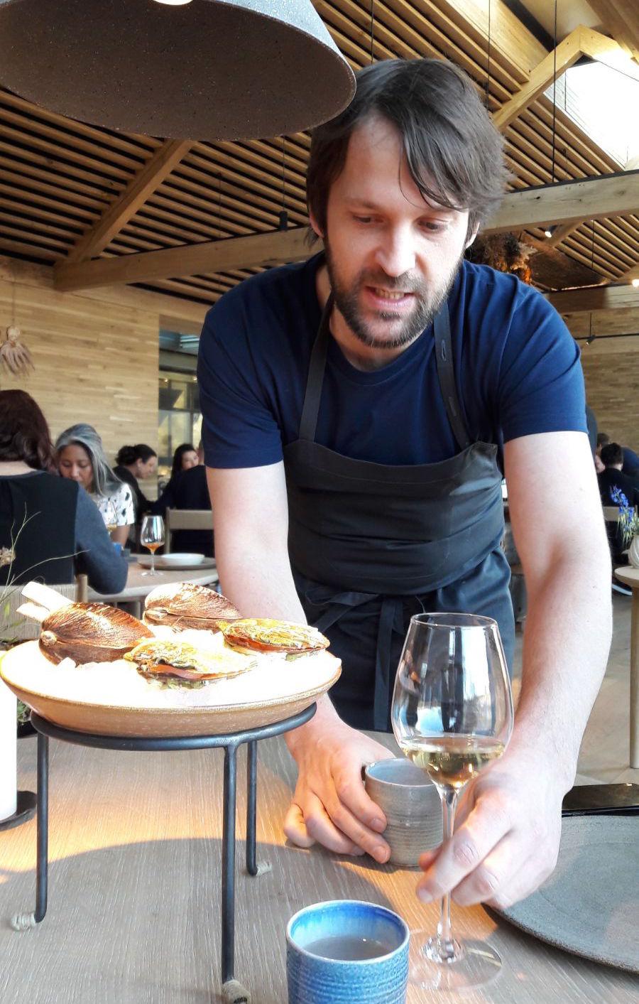 Photogallery






Noma 2.0 is in Christianshavn, the same neighbourhood as the first Noma. They’re 1.5 km apart, a 15 minutes’ walk. This picture shows the distance: to the right, there’s the warehouse of the old location (in Strandgade 93), to the left the smokestacks of Amager Bakke, the futuristic incinerator below which stands the new Noma (in Refshalevej 96)









Weather permitting, you can have a peaceful walk between the previous Noma to today’s Noma 









The new Noma is on an isthmus surrounded by canals. In the future they also want to reclaim some small parts for hydroponic farming









Bucolic scene with incinerator









Bikes left outside Noma. It’s the favourite means of transport among employees and interns









The long path taking you to the restaurant. The fence on the left shows how works are still in progress









On the two sides of the path there are the two new sections of the "Noma village", each with its specialty: grill, hot dishes, snacks, fermentations... In one month’s time, behind the fence we’ll have greenhouses, the test kitchen and the bread making area









To the left, before arriving at the restaurant, the future test kitchen. This will be run by Danish Mette Søberg with Riccardo Canella. "My first challenge?", reveals the latter, for a long time sous chef here, "is to work on the extraction of essential oils" 









The future Test Kitchen









The temporary gangway to the restaurant









Looking from the door to the restaurant, there’s a lake and the smokestacks puffing vapour from the Amager Bakke incinerator. It’s a futuristic project, both in terms of sustainability and architecture. It’s also known as Amager Slope or Copenhill, and its author is starchitect Bjarke Ingels, the same who designed the new Noma. Gradually opened in 2017, it transforms waste into electricity. The goal is to donate electricity to 62500 houses and hot water to 160000. It’s part of an ambitious project to transform Copenhagen into the first city in the world in terms of zero carbon dioxide emissions by 2025. By autumn this year, Amager Slope should open its ski slope, a climbing wall and a hiking track. You’ll be able to slalom on top of the incinerator









The windows from the dining room









Work is still in progress (photo taken on Thursday 22nd February 2018)









Traces of the old building, with barbwire. During WWII, they built bombs here









A view of the dining room at night (photo from Instagram/James Spreadbury)









The entrance to the restaurant









The usual ritual: a welcome from the entire staff, led by Rene Redzepi 









Behind the entrance, there are formaldehyde tanks with creatures from the abyss, shaped as in Damien Hirst’s works. The first image once you’re inside is to reproduce a Scandinavian village









The service kitchen is larger, more spacious and a completely open view from the dining room: "It’s the heart of our Scandinavian village", says Canella. There are 30 cooks working here now. 10 more, compared to the previous Noma









Large skylights above the cooks









There are currently more than 100 people working at Noma, of whom 63 are cooks - 26 employed, and 37 as interns









There’s fire in the kitchen, for the first time









On average, the dining room can host 40 people. "However", complains Redzepi, "there’s always someone who turns up with more people than the ones booked"









While the dining room is set, the outside space still need to be defined









The staff made the dining room decorations. There are no overly expensive designer objects









Dried fish









Seaweeds hanging from the ceiling









Dried octopus heads preserved in salt (with a balloon inside)














Bjarke Ingels and Redzepi’s plan included the wish to recreate a Danish farm









The luminous private dining room









The counter in the dining room. They drew the beams from the canals around the restaurant














The mise en place, bare and strictly without tablecloths









Water jug, candle, flower vase









A warm cloth, to begin with









Australian James Spreadbury, restaurant manager and partner at Noma









Welcome drink: Champagne extra brut Blanc d'Argile from Domaine Vouette et Sorbée









Sea Snail broth. The welcome dish at Noma is, as usual, a hot “soup”









Stefano Ferrano, from Torino, presents the following dish. There’s always a large Italian colony at Noma









Venus clams. Molluscs from Limfjord (Denmark) mixed with decorative clams, typical of Noma. Beside them, shells with blackcurrant oil and “Danish curry”, made with toasted coriander seeds, fennel and fennel seeds









The lid above the following dish. The mussels are not edible









Best of the mussel. Mussels open in a pan with water of fermented barley. «We recreate the mussel», says Canella, «using just the flaps, the best part». The paste is made with scraps from the mussel, cooked in smoked butter









The next dish is conceived and served by Riccardo Canella, the Paduan sous chef who conquered Redzepi, here with dining room intern Feline Hansen









Dried fruits and shrimps. In this case, the heads are not a decoration, but you must suck them –best not eat them because of the bones: they’re filled with a lovely prawn cream. The small crustaceans are still alive when they arrive on a ship straight from the Norwegian fjords









Inside, under the prawn broth skin, there’s fruits and vegetable preserved from the previous summer with raw prawns and gooseberry sauce with blackcurrant oil









How about beverages? There are three options, as usual: wine pairing (6 glasses plus a beer plus sake, 150 euros), juice pairing (juices and extracts, 110 euros) or a choice from the list. They recommend Champagne Chut... Libre from Val'Frison pour Dominique Derain, 50% Pinot Noir, 50% Chardonnay created by a couple who met at the old Noma. Well bodied, it will be a good match throughout the meal









As usual, the wine list starts with Champagne: the cheapest is 1400 crowns (190 euros)









White wines begin with labels from cold climates: first Austria, increasingly popular for its natural wines, then Germany, then all the others









How about Italy? Few names, among the white wines, from North and South









A dozen red/orange wines









Sea star, that is to say Cured trout roe and eggs, the emblem dish of this new era. They recommend moving the teaspoon from the heart to the edges of the tentacles. Aesthetics win over taste









Jellyfish. The jellyfish gel in the middle is in fact a clarified broth of calamari and recalls the texture of jellyfish. "We tried to cook jellyfish in 300 different ways but none was convincing so far". All around, pickled seaweed. The gel is almost tasteless, it serves to clean the palate after the pickles






