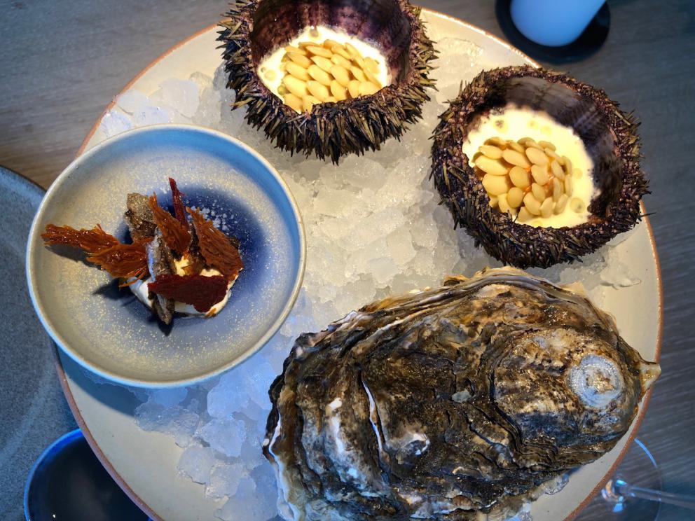 Photogallery






Noma 2.0 is in Christianshavn, the same neighbourhood as the first Noma. They’re 1.5 km apart, a 15 minutes’ walk. This picture shows the distance: to the right, there’s the warehouse of the old location (in Strandgade 93), to the left the smokestacks of Amager Bakke, the futuristic incinerator below which stands the new Noma (in Refshalevej 96)









Weather permitting, you can have a peaceful walk between the previous Noma to today’s Noma 









The new Noma is on an isthmus surrounded by canals. In the future they also want to reclaim some small parts for hydroponic farming









Bucolic scene with incinerator









Bikes left outside Noma. It’s the favourite means of transport among employees and interns









The long path taking you to the restaurant. The fence on the left shows how works are still in progress









On the two sides of the path there are the two new sections of the "Noma village", each with its specialty: grill, hot dishes, snacks, fermentations... In one month’s time, behind the fence we’ll have greenhouses, the test kitchen and the bread making area









To the left, before arriving at the restaurant, the future test kitchen. This will be run by Danish Mette Søberg with Riccardo Canella. "My first challenge?", reveals the latter, for a long time sous chef here, "is to work on the extraction of essential oils" 









The future Test Kitchen









The temporary gangway to the restaurant









Looking from the door to the restaurant, there’s a lake and the smokestacks puffing vapour from the Amager Bakke incinerator. It’s a futuristic project, both in terms of sustainability and architecture. It’s also known as Amager Slope or Copenhill, and its author is starchitect Bjarke Ingels, the same who designed the new Noma. Gradually opened in 2017, it transforms waste into electricity. The goal is to donate electricity to 62500 houses and hot water to 160000. It’s part of an ambitious project to transform Copenhagen into the first city in the world in terms of zero carbon dioxide emissions by 2025. By autumn this year, Amager Slope should open its ski slope, a climbing wall and a hiking track. You’ll be able to slalom on top of the incinerator









The windows from the dining room









Work is still in progress (photo taken on Thursday 22nd February 2018)









Traces of the old building, with barbwire. During WWII, they built bombs here









A view of the dining room at night (photo from Instagram/James Spreadbury)









The entrance to the restaurant









The usual ritual: a welcome from the entire staff, led by Rene Redzepi 









Behind the entrance, there are formaldehyde tanks with creatures from the abyss, shaped as in Damien Hirst’s works. The first image once you’re inside is to reproduce a Scandinavian village









The service kitchen is larger, more spacious and a completely open view from the dining room: "It’s the heart of our Scandinavian village", says Canella. There are 30 cooks working here now. 10 more, compared to the previous Noma









Large skylights above the cooks









There are currently more than 100 people working at Noma, of whom 63 are cooks - 26 employed, and 37 as interns









There’s fire in the kitchen, for the first time









On average, the dining room can host 40 people. "However", complains Redzepi, "there’s always someone who turns up with more people than the ones booked"









While the dining room is set, the outside space still need to be defined









The staff made the dining room decorations. There are no overly expensive designer objects









Dried fish









Seaweeds hanging from the ceiling









Dried octopus heads preserved in salt (with a balloon inside)














Bjarke Ingels and Redzepi’s plan included the wish to recreate a Danish farm









The luminous private dining room









The counter in the dining room. They drew the beams from the canals around the restaurant














The mise en place, bare and strictly without tablecloths









Water jug, candle, flower vase









A warm cloth, to begin with









Australian James Spreadbury, restaurant manager and partner at Noma









Welcome drink: Champagne extra brut Blanc d'Argile from Domaine Vouette et Sorbée









Sea Snail broth. The welcome dish at Noma is, as usual, a hot “soup”









Stefano Ferrano, from Torino, presents the following dish. There’s always a large Italian colony at Noma









Venus clams. Molluscs from Limfjord (Denmark) mixed with decorative clams, typical of Noma. Beside them, shells with blackcurrant oil and “Danish curry”, made with toasted coriander seeds, fennel and fennel seeds









The lid above the following dish. The mussels are not edible









Best of the mussel. Mussels open in a pan with water of fermented barley. «We recreate the mussel», says Canella, «using just the flaps, the best part». The paste is made with scraps from the mussel, cooked in smoked butter









The next dish is conceived and served by Riccardo Canella, the Paduan sous chef who conquered Redzepi, here with dining room intern Feline Hansen









Dried fruits and shrimps. In this case, the heads are not a decoration, but you must suck them –best not eat them because of the bones: they’re filled with a lovely prawn cream. The small crustaceans are still alive when they arrive on a ship straight from the Norwegian fjords









Inside, under the prawn broth skin, there’s fruits and vegetable preserved from the previous summer with raw prawns and gooseberry sauce with blackcurrant oil









How about beverages? There are three options, as usual: wine pairing (6 glasses plus a beer plus sake, 150 euros), juice pairing (juices and extracts, 110 euros) or a choice from the list. They recommend Champagne Chut... Libre from Val'Frison pour Dominique Derain, 50% Pinot Noir, 50% Chardonnay created by a couple who met at the old Noma. Well bodied, it will be a good match throughout the meal









As usual, the wine list starts with Champagne: the cheapest is 1400 crowns (190 euros)









White wines begin with labels from cold climates: first Austria, increasingly popular for its natural wines, then Germany, then all the others









How about Italy? Few names, among the white wines, from North and South









A dozen red/orange wines









Sea star, that is to say Cured trout roe and eggs, the emblem dish of this new era. They recommend moving the teaspoon from the heart to the edges of the tentacles. Aesthetics win over taste









Jellyfish. The jellyfish gel in the middle is in fact a clarified broth of calamari and recalls the texture of jellyfish. "We tried to cook jellyfish in 300 different ways but none was convincing so far". All around, pickled seaweed. The gel is almost tasteless, it serves to clean the palate after the pickles









Rene Redzepi himself presents the content of the first Seafood platter.









French style service with a small plate below for the empty shells









It’s made of two (per two) pieces: queen clam and mahogany clam









The Queen Clam is a very small variety of Norwegian scallop, seasoned with a vegetal reduction made with cucumber and peas miso. The red tip is the coral. "You should eat it using the top shell ", recommends Redzepi









The Mahogany clam is a centennial clam from the North Sea, very popular in Denmark and Iceland. «You can tell the age by the number of rings, like with trees», says Redzepi, «In 2007 they found one that was 450 years old. We’ve had it in the menu for a decade or so». The razor clam shell serves as teaspoon









Inside, there’s obviously not just a bare Mahogany. «We open it when the client arrives», says Canella, «remove the entrails and then slice it so as to preserve its firm texture». It’s seasoned with stir-fried gooseberry, capers of blackcurrants and blackcurrant sprouts preserved in vinegar. The sauce is made with mussel water









A note on the chairs: they’re more minimalistic. They’re no longer covered in sheepskin or leather from other Danish animals, in the style of Noma 1.0. The most significant element is the napkin: if you go to the toilet, they no longer replace it with a new one. There’s the old one, tied to the chair. "This is sustainability", they explain






