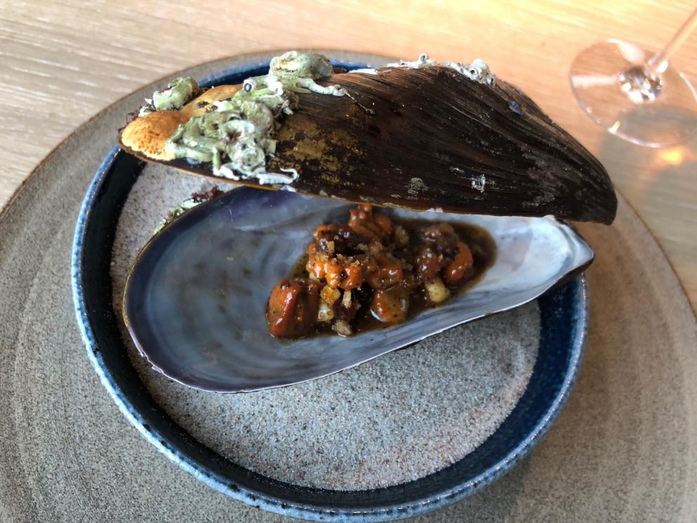 Photogallery






Noma 2.0 is in Christianshavn, the same neighbourhood as the first Noma. They’re 1.5 km apart, a 15 minutes’ walk. This picture shows the distance: to the right, there’s the warehouse of the old location (in Strandgade 93), to the left the smokestacks of Amager Bakke, the futuristic incinerator below which stands the new Noma (in Refshalevej 96)









Weather permitting, you can have a peaceful walk between the previous Noma to today’s Noma 









The new Noma is on an isthmus surrounded by canals. In the future they also want to reclaim some small parts for hydroponic farming









Bucolic scene with incinerator









Bikes left outside Noma. It’s the favourite means of transport among employees and interns









The long path taking you to the restaurant. The fence on the left shows how works are still in progress









On the two sides of the path there are the two new sections of the "Noma village", each with its specialty: grill, hot dishes, snacks, fermentations... In one month’s time, behind the fence we’ll have greenhouses, the test kitchen and the bread making area









To the left, before arriving at the restaurant, the future test kitchen. This will be run by Danish Mette Søberg with Riccardo Canella. "My first challenge?", reveals the latter, for a long time sous chef here, "is to work on the extraction of essential oils" 









The future Test Kitchen









The temporary gangway to the restaurant









Looking from the door to the restaurant, there’s a lake and the smokestacks puffing vapour from the Amager Bakke incinerator. It’s a futuristic project, both in terms of sustainability and architecture. It’s also known as Amager Slope or Copenhill, and its author is starchitect Bjarke Ingels, the same who designed the new Noma. Gradually opened in 2017, it transforms waste into electricity. The goal is to donate electricity to 62500 houses and hot water to 160000. It’s part of an ambitious project to transform Copenhagen into the first city in the world in terms of zero carbon dioxide emissions by 2025. By autumn this year, Amager Slope should open its ski slope, a climbing wall and a hiking track. You’ll be able to slalom on top of the incinerator









The windows from the dining room









Work is still in progress (photo taken on Thursday 22nd February 2018)









Traces of the old building, with barbwire. During WWII, they built bombs here









A view of the dining room at night (photo from Instagram/James Spreadbury)









The entrance to the restaurant









The usual ritual: a welcome from the entire staff, led by Rene Redzepi 









Behind the entrance, there are formaldehyde tanks with creatures from the abyss, shaped as in Damien Hirst’s works. The first image once you’re inside is to reproduce a Scandinavian village









The service kitchen is larger, more spacious and a completely open view from the dining room: "It’s the heart of our Scandinavian village", says Canella. There are 30 cooks working here now. 10 more, compared to the previous Noma









Large skylights above the cooks









There are currently more than 100 people working at Noma, of whom 63 are cooks - 26 employed, and 37 as interns









There’s fire in the kitchen, for the first time









On average, the dining room can host 40 people. "However", complains Redzepi, "there’s always someone who turns up with more people than the ones booked"









While the dining room is set, the outside space still need to be defined









The staff made the dining room decorations. There are no overly expensive designer objects









Dried fish









Seaweeds hanging from the ceiling









Dried octopus heads preserved in salt (with a balloon inside)














Bjarke Ingels and Redzepi’s plan included the wish to recreate a Danish farm









The luminous private dining room









The counter in the dining room. They drew the beams from the canals around the restaurant














The mise en place, bare and strictly without tablecloths









Water jug, candle, flower vase









A warm cloth, to begin with









Australian James Spreadbury, restaurant manager and partner at Noma









Welcome drink: Champagne extra brut Blanc d'Argile from Domaine Vouette et Sorbée









Sea Snail broth. The welcome dish at Noma is, as usual, a hot “soup”









Stefano Ferrano, from Torino, presents the following dish. There’s always a large Italian colony at Noma









Venus clams. Molluscs from Limfjord (Denmark) mixed with decorative clams, typical of Noma. Beside them, shells with blackcurrant oil and “Danish curry”, made with toasted coriander seeds, fennel and fennel seeds









The lid above the following dish. The mussels are not edible









Best of the mussel. Mussels open in a pan with water of fermented barley. «We recreate the mussel», says Canella, «using just the flaps, the best part». The paste is made with scraps from the mussel, cooked in smoked butter









The next dish is conceived and served by Riccardo Canella, the Paduan sous chef who conquered Redzepi, here with dining room intern Feline Hansen









Dried fruits and shrimps. In this case, the heads are not a decoration, but you must suck them –best not eat them because of the bones: they’re filled with a lovely prawn cream. The small crustaceans are still alive when they arrive on a ship straight from the Norwegian fjords









Inside, under the prawn broth skin, there’s fruits and vegetable preserved from the previous summer with raw prawns and gooseberry sauce with blackcurrant oil









How about beverages? There are three options, as usual: wine pairing (6 glasses plus a beer plus sake, 150 euros), juice pairing (juices and extracts, 110 euros) or a choice from the list. They recommend Champagne Chut... Libre from Val'Frison pour Dominique Derain, 50% Pinot Noir, 50% Chardonnay created by a couple who met at the old Noma. Well bodied, it will be a good match throughout the meal









As usual, the wine list starts with Champagne: the cheapest is 1400 crowns (190 euros)









White wines begin with labels from cold climates: first Austria, increasingly popular for its natural wines, then Germany, then all the others









How about Italy? Few names, among the white wines, from North and South









A dozen red/orange wines









Sea star, that is to say Cured trout roe and eggs, the emblem dish of this new era. They recommend moving the teaspoon from the heart to the edges of the tentacles. Aesthetics win over taste









Jellyfish. The jellyfish gel in the middle is in fact a clarified broth of calamari and recalls the texture of jellyfish. "We tried to cook jellyfish in 300 different ways but none was convincing so far". All around, pickled seaweed. The gel is almost tasteless, it serves to clean the palate after the pickles









Rene Redzepi himself presents the content of the first Seafood platter.









French style service with a small plate below for the empty shells









It’s made of two (per two) pieces: queen clam and mahogany clam









The Queen Clam is a very small variety of Norwegian scallop, seasoned with a vegetal reduction made with cucumber and peas miso. The red tip is the coral. "You should eat it using the top shell ", recommends Redzepi









The Mahogany clam is a centennial clam from the North Sea, very popular in Denmark and Iceland. «You can tell the age by the number of rings, like with trees», says Redzepi, «In 2007 they found one that was 450 years old. We’ve had it in the menu for a decade or so». The razor clam shell serves as teaspoon









Inside, there’s obviously not just a bare Mahogany. «We open it when the client arrives», says Canella, «remove the entrails and then slice it so as to preserve its firm texture». It’s seasoned with stir-fried gooseberry, capers of blackcurrants and blackcurrant sprouts preserved in vinegar. The sauce is made with mussel water









A note on the chairs: they’re more minimalistic. They’re no longer covered in sheepskin or leather from other Danish animals, in the style of Noma 1.0. The most significant element is the napkin: if you go to the toilet, they no longer replace it with a new one. There’s the old one, tied to the chair. "This is sustainability", they explain









The second seafood platter. It’s made of Sea cucumber and sour cream in various textures, including «chicharrones» (at 9 o’clock), sea urchins (at 12 and 3) and gigas oyster (at 6)









Sea urchins pressed in fresh cream and pumpkin seeds









Giga Oyster, a huge oyster from the Pacific, quickly steamed and garnished with pickles. Noma only reserved one thousand of these. In the menu until available






