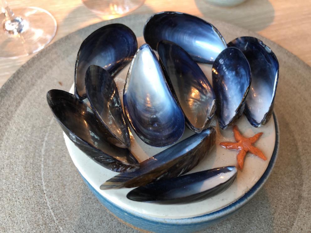 Photogallery






Noma 2.0 is in Christianshavn, the same neighbourhood as the first Noma. They’re 1.5 km apart, a 15 minutes’ walk. This picture shows the distance: to the right, there’s the warehouse of the old location (in Strandgade 93), to the left the smokestacks of Amager Bakke, the futuristic incinerator below which stands the new Noma (in Refshalevej 96)









Weather permitting, you can have a peaceful walk between the previous Noma to today’s Noma 









The new Noma is on an isthmus surrounded by canals. In the future they also want to reclaim some small parts for hydroponic farming









Bucolic scene with incinerator









Bikes left outside Noma. It’s the favourite means of transport among employees and interns









The long path taking you to the restaurant. The fence on the left shows how works are still in progress









On the two sides of the path there are the two new sections of the "Noma village", each with its specialty: grill, hot dishes, snacks, fermentations... In one month’s time, behind the fence we’ll have greenhouses, the test kitchen and the bread making area









To the left, before arriving at the restaurant, the future test kitchen. This will be run by Danish Mette Søberg with Riccardo Canella. "My first challenge?", reveals the latter, for a long time sous chef here, "is to work on the extraction of essential oils" 









The future Test Kitchen









The temporary gangway to the restaurant









Looking from the door to the restaurant, there’s a lake and the smokestacks puffing vapour from the Amager Bakke incinerator. It’s a futuristic project, both in terms of sustainability and architecture. It’s also known as Amager Slope or Copenhill, and its author is starchitect Bjarke Ingels, the same who designed the new Noma. Gradually opened in 2017, it transforms waste into electricity. The goal is to donate electricity to 62500 houses and hot water to 160000. It’s part of an ambitious project to transform Copenhagen into the first city in the world in terms of zero carbon dioxide emissions by 2025. By autumn this year, Amager Slope should open its ski slope, a climbing wall and a hiking track. You’ll be able to slalom on top of the incinerator









The windows from the dining room









Work is still in progress (photo taken on Thursday 22nd February 2018)









Traces of the old building, with barbwire. During WWII, they built bombs here









A view of the dining room at night (photo from Instagram/James Spreadbury)









The entrance to the restaurant









The usual ritual: a welcome from the entire staff, led by Rene Redzepi 









Behind the entrance, there are formaldehyde tanks with creatures from the abyss, shaped as in Damien Hirst’s works. The first image once you’re inside is to reproduce a Scandinavian village









The service kitchen is larger, more spacious and a completely open view from the dining room: "It’s the heart of our Scandinavian village", says Canella. There are 30 cooks working here now. 10 more, compared to the previous Noma









Large skylights above the cooks









There are currently more than 100 people working at Noma, of whom 63 are cooks - 26 employed, and 37 as interns









There’s fire in the kitchen, for the first time









On average, the dining room can host 40 people. "However", complains Redzepi, "there’s always someone who turns up with more people than the ones booked"









While the dining room is set, the outside space still need to be defined









The staff made the dining room decorations. There are no overly expensive designer objects









Dried fish









Seaweeds hanging from the ceiling









Dried octopus heads preserved in salt (with a balloon inside)














Bjarke Ingels and Redzepi’s plan included the wish to recreate a Danish farm









The luminous private dining room









The counter in the dining room. They drew the beams from the canals around the restaurant














The mise en place, bare and strictly without tablecloths









Water jug, candle, flower vase









A warm cloth, to begin with









Australian James Spreadbury, restaurant manager and partner at Noma









Welcome drink: Champagne extra brut Blanc d'Argile from Domaine Vouette et Sorbée









Sea Snail broth. The welcome dish at Noma is, as usual, a hot “soup”









Stefano Ferrano, from Torino, presents the following dish. There’s always a large Italian colony at Noma









Venus clams. Molluscs from Limfjord (Denmark) mixed with decorative clams, typical of Noma. Beside them, shells with blackcurrant oil and “Danish curry”, made with toasted coriander seeds, fennel and fennel seeds






