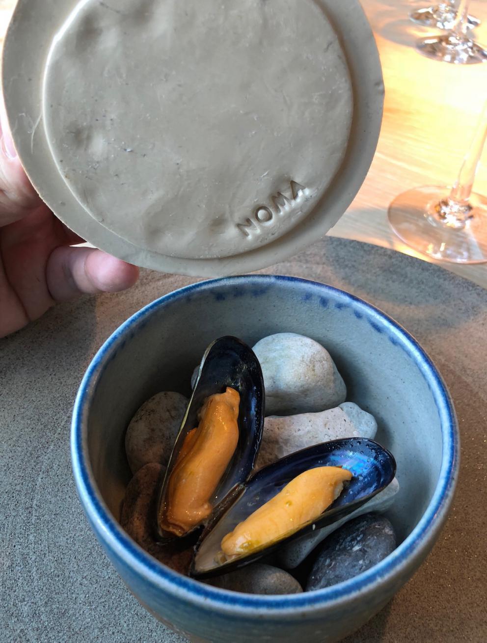 Photogallery






Noma 2.0 is in Christianshavn, the same neighbourhood as the first Noma. They’re 1.5 km apart, a 15 minutes’ walk. This picture shows the distance: to the right, there’s the warehouse of the old location (in Strandgade 93), to the left the smokestacks of Amager Bakke, the futuristic incinerator below which stands the new Noma (in Refshalevej 96)









Weather permitting, you can have a peaceful walk between the previous Noma to today’s Noma 









The new Noma is on an isthmus surrounded by canals. In the future they also want to reclaim some small parts for hydroponic farming









Bucolic scene with incinerator









Bikes left outside Noma. It’s the favourite means of transport among employees and interns









The long path taking you to the restaurant. The fence on the left shows how works are still in progress









On the two sides of the path there are the two new sections of the "Noma village", each with its specialty: grill, hot dishes, snacks, fermentations... In one month’s time, behind the fence we’ll have greenhouses, the test kitchen and the bread making area









To the left, before arriving at the restaurant, the future test kitchen. This will be run by Danish Mette Søberg with Riccardo Canella. "My first challenge?", reveals the latter, for a long time sous chef here, "is to work on the extraction of essential oils" 









The future Test Kitchen









The temporary gangway to the restaurant









Looking from the door to the restaurant, there’s a lake and the smokestacks puffing vapour from the Amager Bakke incinerator. It’s a futuristic project, both in terms of sustainability and architecture. It’s also known as Amager Slope or Copenhill, and its author is starchitect Bjarke Ingels, the same who designed the new Noma. Gradually opened in 2017, it transforms waste into electricity. The goal is to donate electricity to 62500 houses and hot water to 160000. It’s part of an ambitious project to transform Copenhagen into the first city in the world in terms of zero carbon dioxide emissions by 2025. By autumn this year, Amager Slope should open its ski slope, a climbing wall and a hiking track. You’ll be able to slalom on top of the incinerator









The windows from the dining room









Work is still in progress (photo taken on Thursday 22nd February 2018)









Traces of the old building, with barbwire. During WWII, they built bombs here









A view of the dining room at night (photo from Instagram/James Spreadbury)









The entrance to the restaurant









The usual ritual: a welcome from the entire staff, led by Rene Redzepi 









Behind the entrance, there are formaldehyde tanks with creatures from the abyss, shaped as in Damien Hirst’s works. The first image once you’re inside is to reproduce a Scandinavian village









The service kitchen is larger, more spacious and a completely open view from the dining room: "It’s the heart of our Scandinavian village", says Canella. There are 30 cooks working here now. 10 more, compared to the previous Noma









Large skylights above the cooks









There are currently more than 100 people working at Noma, of whom 63 are cooks - 26 employed, and 37 as interns









There’s fire in the kitchen, for the first time









On average, the dining room can host 40 people. "However", complains Redzepi, "there’s always someone who turns up with more people than the ones booked"









While the dining room is set, the outside space still need to be defined









The staff made the dining room decorations. There are no overly expensive designer objects









Dried fish









Seaweeds hanging from the ceiling









Dried octopus heads preserved in salt (with a balloon inside)














Bjarke Ingels and Redzepi’s plan included the wish to recreate a Danish farm









The luminous private dining room









The counter in the dining room. They drew the beams from the canals around the restaurant














The mise en place, bare and strictly without tablecloths









Water jug, candle, flower vase









A warm cloth, to begin with









Australian James Spreadbury, restaurant manager and partner at Noma









Welcome drink: Champagne extra brut Blanc d'Argile from Domaine Vouette et Sorbée









Sea Snail broth. The welcome dish at Noma is, as usual, a hot “soup”









Stefano Ferrano, from Torino, presents the following dish. There’s always a large Italian colony at Noma









Venus clams. Molluscs from Limfjord (Denmark) mixed with decorative clams, typical of Noma. Beside them, shells with blackcurrant oil and “Danish curry”, made with toasted coriander seeds, fennel and fennel seeds









The lid above the following dish. The mussels are not edible






