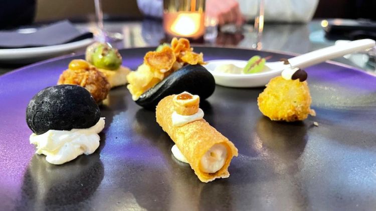 Stuzzichini di benvenuto: Bao al nero di seppia con calamaretti fritti e maionese al lime, Ghianda di quaglia con il suo bagnetto, Sassolino del Meta con cremoso di formaggio al tartufo, Crocchetta di baccalà con maionese all’aglio, Crocchetta di Patanegra con senape al miele, Oliva di foie gras e Sauternes, Cannoncino di Jersey Blue e nocciola del Piemonte, Panna cotta ai carciofi e ostrica
