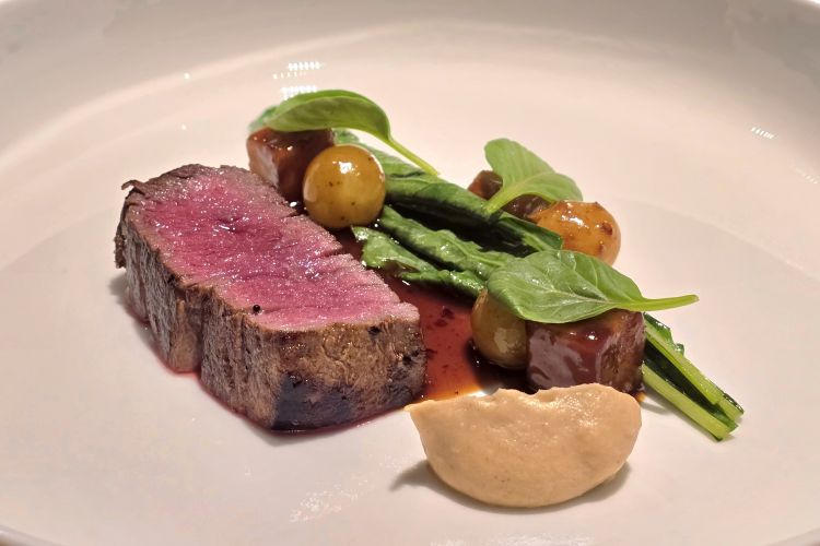 Manzo con quenelle di sedano rapa, foglie di cavolo nero e uva alla brace. Ogni elemento subisce la carezza della brace, che dona un filo conduttore aromatico e avvolgente. Piatto di grande solidità, dove dolcezze, note vegetali e umami si intrecciano in armonia rigorosa
