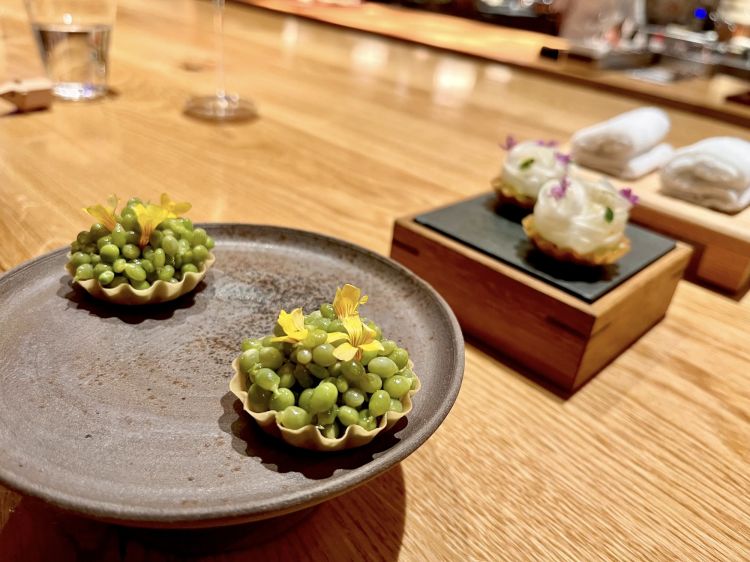 Tartelletta di piselli lagrima, un simbolo commestibile dei Paesi Baschi: è una tartelletta wonton con piselli grigliati, conditi con olio d'erba cipollina, e sgombro marinato in fondo. Sullo sfondo, si intravede un'altra Tartelletta di pasta di gyoza con astice in salsa salpicon (simile alla salsa verde) con asparagi bianchi
