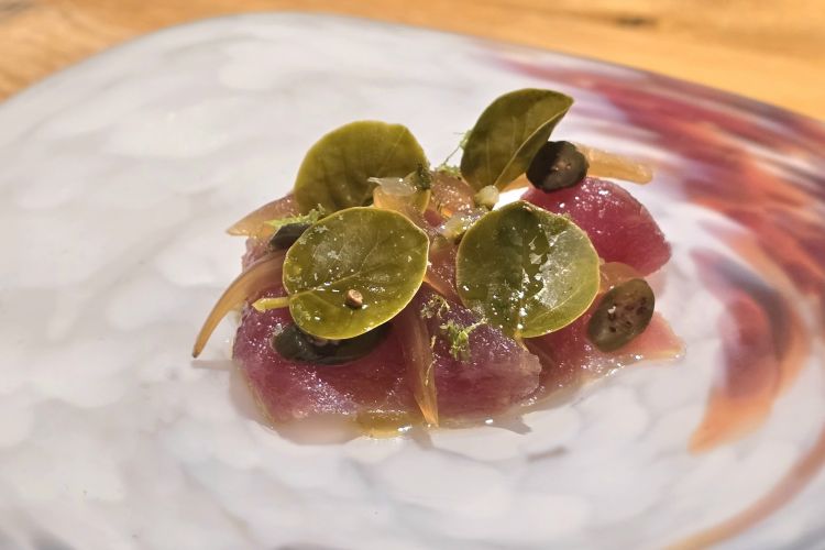 Carpaccio di tonno con foglie di cappero e cucunci, yuzu di Noto sottaceto e cipolla giarratana, prima cotta alla brace poi passata in aceto. Un antipasto che celebra mare e terra di Sicilia, giocato su un equilibrio armonico tra spigoli acetici e freschezza agrumata
