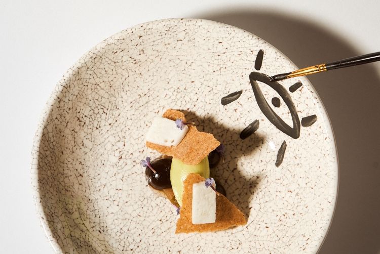 Apotropaico: ruta, aglio nero, fico in conserva, “ossa dei morti” con rosa canina e idromele montato. «Nel Mediterraneo l’autunno è tempo di passaggi: la fine dei raccolti, l’accorciarsi della luce, l’emergere della memoria dei morti. È anche la stagione in cui si intensificano pratiche di protezione contro il male invisibile, che nelle culture locali ha preso la forma del malocchio: fascinum per i Romani, jettatura in area napoletana, baskanía in greco, nazar in turco, ʿayn al-ḥasad in arabo. Ernesto De Martino, in Sud e Magia, ha mostrato come il malocchio rappresenti un dispositivo culturale che traduce vulnerabilità e invidia in un linguaggio simbolico. Non si tratta di semplice superstizione: lo “sguardo che nuoce” è un modo di dare forma a tensioni sociali, a paure di perdita, a crisi della “presenza”. Uno sguardo capace di sottrarre forza vitale e destabilizzare l’individuo e la comunità. Difendersi significava allora dotarsi di un lessico materiale e rituale che andava dal cibo agli scongiuri fino ai gesti delle mani. Si appendevano trecce d’aglio alle porte: se col tempo annerivano, significava che avevano assorbito il malocchio. E ancora amuleti in corallo, gesti come la “fica” o le corna, pani incisi: il Mediterraneo ha costruito una vera grammatica della prevenzione, in cui anche il cibo partecipa come barriera simbolica. In questa logica l’apotropaico non elimina il pericolo, ma lo devia, lo tiene a distanza, consente alla comunità di abitare la propria fragilità senza soccombere»