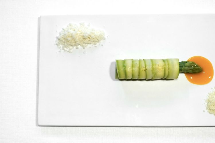 Asparagus with elderberry flower and macadamia nut: the stripe of green asparagus is wrapped around a sorbet of white asparagus. On top, the tip of a green asparagus placed on mandarin juice. Two powders on the side: on top, macadamia, below, a frozen powder of elderberry flower
