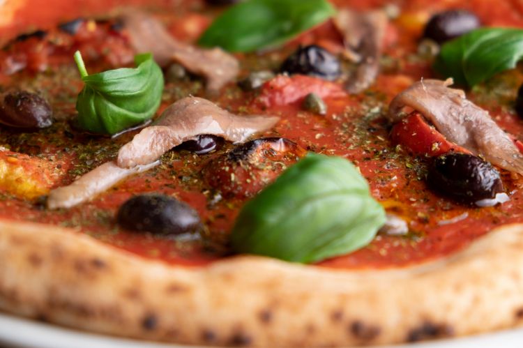 La pizza Piennolo del Vesuvio Dop: pomodoro, acciughe del Mar Cantabrico, olive nere denocciolate cotte al forno, capperi di Pantelleria, origano, olio infuso all’aglio.
