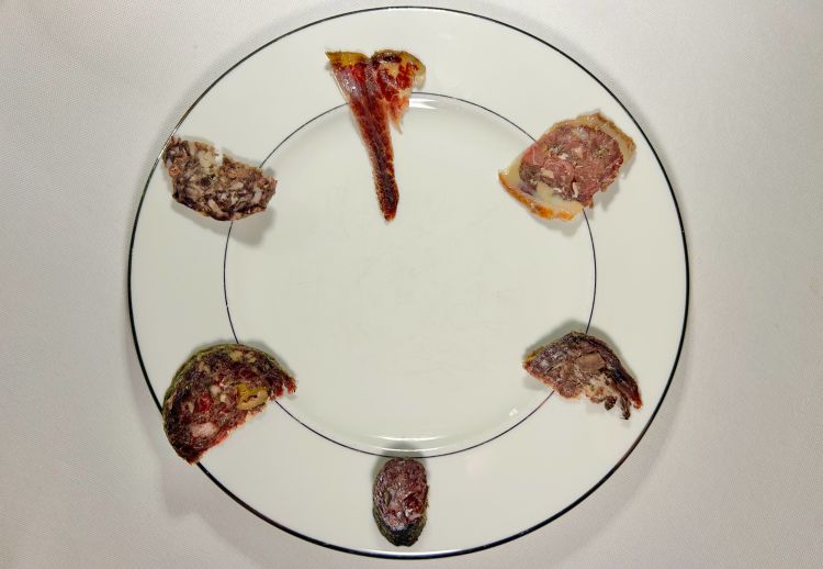 Insaccati. La materia prima proviene da caccia di selezione, con abbattimenti eseguiti da cacciatori certificati e con tracciabilità garantita dall’Asl. A fianco dei salumi viene servito anche un panino preparato con farina di ghiande, cotto a vapore e farcito con carne di recupero di capriolo, in particolare costole e collo (nonché delle verdure in giardiniera). Le fette: 1 salume di colombaccio aromatizzato con ginepro e limone fermentato; 2 salame di capriolo al polline e capperi; 3 salame di palacone di daino, cioè il maschio adulto, servito con olive, cavolo nero stufato e scorza di mandarino (l’idea è abbinare una carne “virile” a note amare e complesse); 4 fesa di daino femmina abbinata a una quantità molto importante di lamponi disidratati, pari al 16%, e bergamotto (la dolcezza e l’acidità del frutto servono a equilibrare il carattere della carne); 5 fesa di cavallo di 14 anni (la carne, macellata nel 2022, non viene salata direttamente, ma immersa in un garum di agnello. In questo modo si ricerca una bassa salinità accompagnata da un’elevata attività enzimatica. In bocca la carne evolve progressivamente, sviluppando con la sua marezzatura anche note vegetali e marine); 6 anatra e cinghiale nella pelle dell’anatra, per mantenere l’umidità («È una sorta di “chimera”, cioè un impasto di carni miste concepito per bilanciare le rispettive componenti grasse, come si usava una volta per i maiali - dice lo chef - Oggi abbiamo cinghiali molto più grassi e quindi vi abbino l’anatra, per ottenere eleganza». La carne viene lavorata con miele di castagno che fa da conservante, lega le carni e evolve, fino all’acidità. C’è un po’ di scorza d’arancia e pochissimo finocchietto selvatico. Il cinghiale proviene da caccia d’appostamento e non da battuta: questo permette di ottenere una carne meno segnata dall’adrenalina dell’animale)