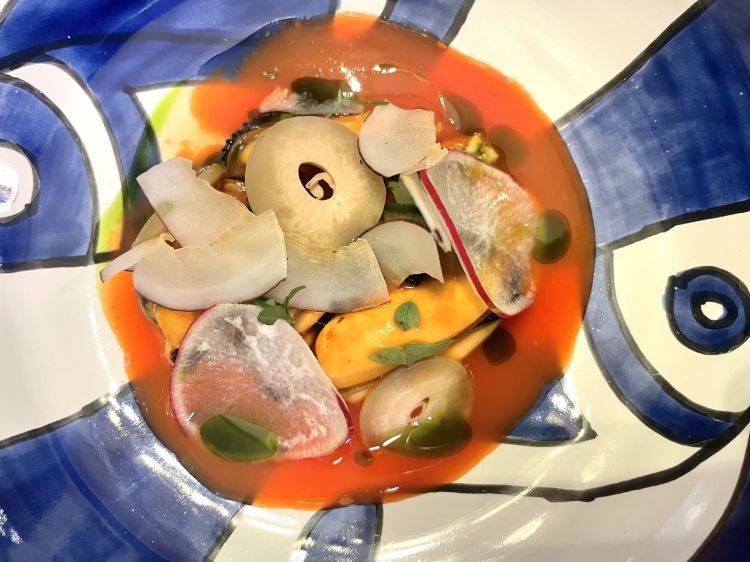 Zuppa di cozze, pomodoro estivo, cocco, facussa e ravanelli
Un piatto davvero complesso, in questo caso meno immediato, ma che ci aiuta a immaginare la cozza in maniera completamente diversa rispetto alla sua preparazione canonica. È una cozza sarda che viene lasciata schiudere e poi passata per pochi minuti alla brace, giusto il tempo di raccogliere nella sua polpa un fumo docile – non di più. Alla base c’è della facussa fermentata – una varietà di melone, molto simile a un cetriolo – e del pomodoro, il ricordo di una conserva estiva, del frutto al naturale, scaldato dal sole, e che quindi trattiene un sapore acidulo, mentre di dissolve la dolcezza; non viene aggiunto sale, ma solo del coriandolo, peschiole - dai tratti acerbi -, caviale, ravanelli e cocco secco, che non rilascia quella parte tostata quasi di frutta secca, ma che vira, invece, verso un’acidità quasi balsamica, resinosa. Il mare inizia a silenziarsi e vengono evocati sentori più terrosi
