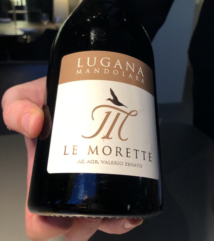 Lugana DOP Mandolara 2021, Le Morette
Al calice, apre la degustazione il Lugana DOP Mandolara 2021 – Le Morette, azienda d’oltre 60 anni, giunta alla terza generazione, a Peschiera del Garda. Un vino che esprime a pieno la tipicità delle uve Turbiana, a partire dal colore, un giallo paglierino con riflessi verdognoli, fino alle sue note spiccate di fiori bianchi che, dopo un breve affinamento in bottiglia, divengono fruttate. Sapore fresco, particolarmente delicato, con un ricorrente sentore di mandorla nel finale, supportato dalla naturale sapidità che origina dai terreni argillosi. Un sorso elegante e una mineralità che attraversa e sostiene i sapori decisi di inizio percorso
