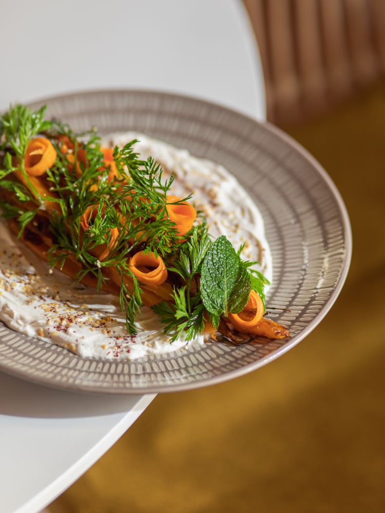 Insalata autunnale marocchina di carote al forno su base di yogurt, tahina e polvere di sumak, condita con vinaigrette di cannella, olio e aceto. Viene servita con uvetta, menta, sesamo e riccioli di carote marinate
