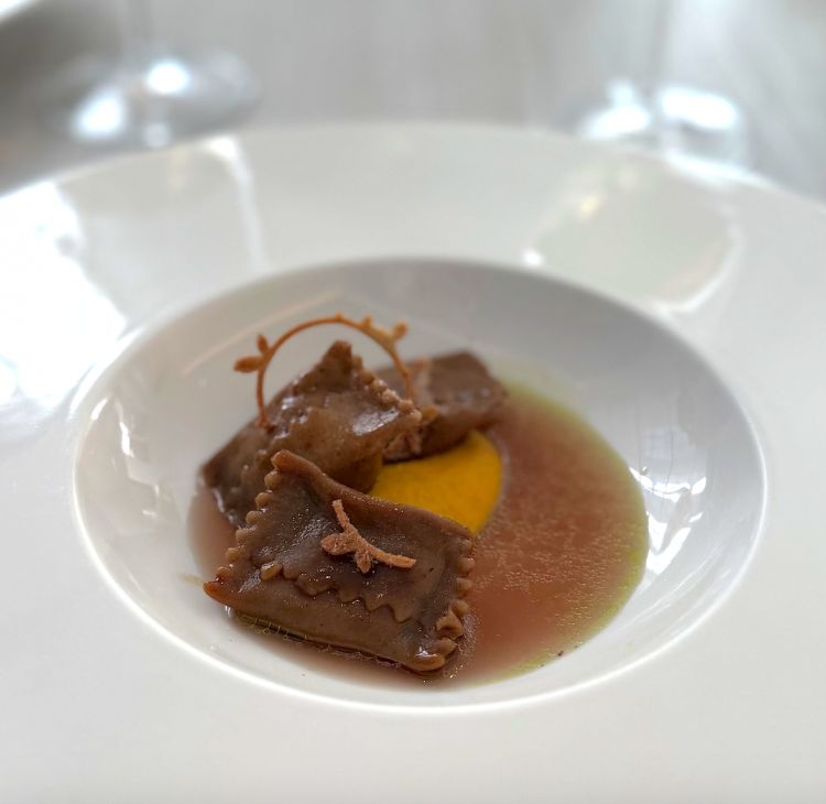 Secondo round di primi con Agnolotti di farina di castagne ripieni di sfilacci di manzo con zafferano, alloro e un consomm&eacute; di rose. Fuori menu, lo chef propone questi agnolotti di farina di castagne&nbsp;serviti con crema allo zafferano, tuille all&rsquo;alloro e polvere di rose, accompagnati da un consomm&eacute; di manzo infuso con rose e alloro. Il ripieno &egrave; intenso e gustoso, cos&igrave; come la salsa e il brodo, profondo e persistente. La pasta, leggermente spessa, limita in parte la finezza del boccone; le note floreali risultano pi&ugrave; accennate che protagoniste. Nel complesso, un piatto saporito e strutturato. In abbinamento,&nbsp;Ros&eacute; Syrah BIO Toscana IGT "Rosa del Castagno" Cantina Fabrizio Dionisio 2022
