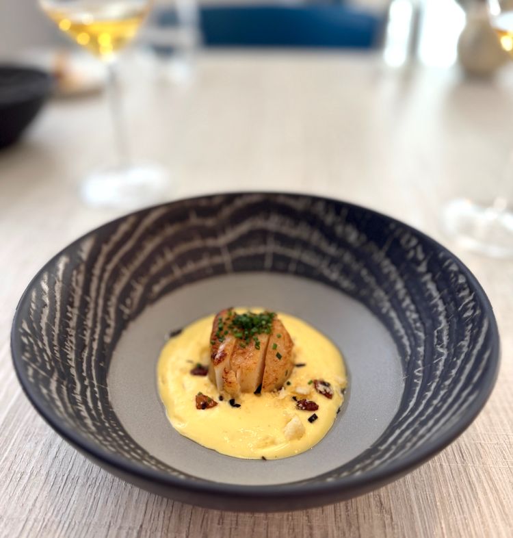 Si procede con Capasanta con spuma alla carbonara, erba cipollina e alga Kombu. La capasanta &egrave; abbinata a una spuma &ldquo;alla carbonara&rdquo; sifonata: al posto del Pecorino Romano, viene usato un Asiago Riserva, pi&ugrave; misurato ma comunque sapido cos&igrave; da non sovrastare la delicatezza del mollusco.&nbsp;Questa volta ci viene servito un calice di Malvasia del Carso DOC Azienda Agricola Kocjancic Rado 2023
