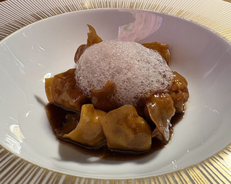 Raviolo - Tempeh, vino rosso e spezie
«E' un tortellino ripieno di tempeh, limone, erbe e pepe. Poi è glassato con un fondo bruno di topinambur, cipolla, zenzero e carote. La parte che abbiamo studiato di più in realtà è la glassa, il fondo bruno. Lo andiamo a fare proprio come un fondo bruno, ma senza ossa, è completamente vegetale, mentre la spuma è fatta con vino, burro e spezie»