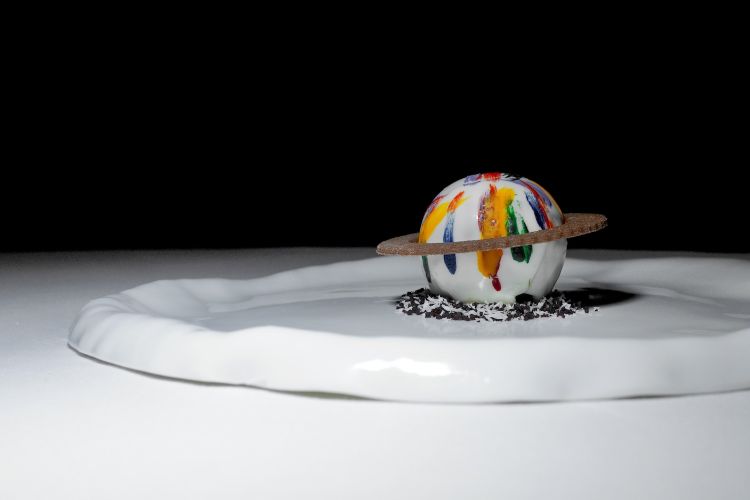 Cioccolato, frutto della passione e vaniglia. La sfera di cioccolato bianco con i colori della bandiera della pace racchiude mousse al cioccolato e una salsa liquida di passion fruit
