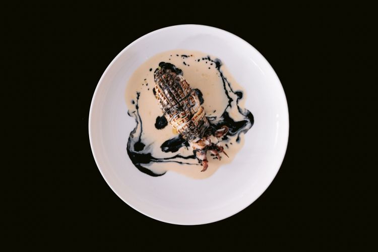Seppia cotta su carbone di legno di ulivo, flambata sulla parte superiore, ripiena con “caponata bianca” di carciofi, capperi e mandorle, guarnita con salsa all’acciuga, sale di Mothia, latte di mandorla e nero di seppia