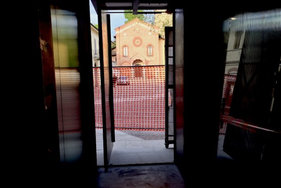 From inside the building site of the new D'O, right where there will be the crystal door, the telephone’s lens pointed at sunset on Friday 15th April 2016 on the facade of the Chiesa Vecchia di San Pietro. The historic elm tree is in the middle of the square, between the restaurant and the place of worship
