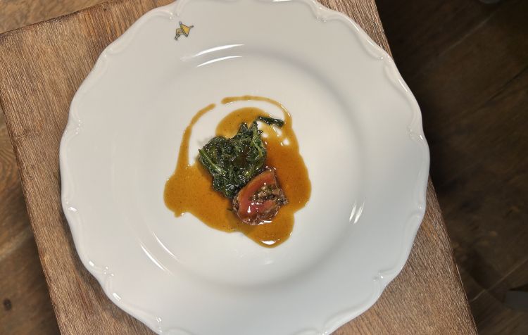 Piccione appeso tutto intero sulle braci per sigillare le carni; poi il petto viene farcito con il suo umido, chiuso nella rete di maiale e cucinato con spezie e odori, poi servito con fondo di piccione e spinacini saltati
