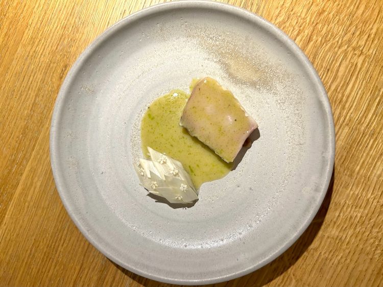 Ricciola frollata una settimana, cotta sulla brace dalla parte della pelle, riposa in letto di alga kombu (è la tecnica giapponese detta kombujime) per acquisire la parte sapida e marina, poi crema di limone fermentato, asparago bianco (sia affumicato sia a crudo), beurre blanc con miso di ceci e kombucha ai fiori di sambuco
